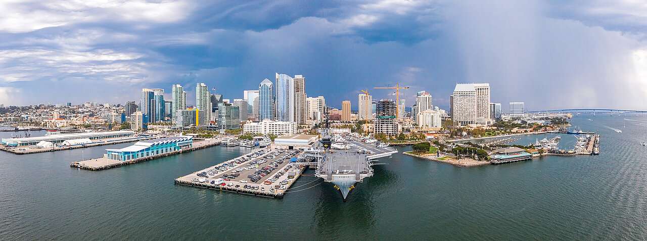 Erstaunlicher Panoramablick auf die Innenstadt von San Diego am Hafen mit vielen Wolkenkratzern und einem riesigen Flugzeugträger, der von der Anlegestelle angedockt wird.