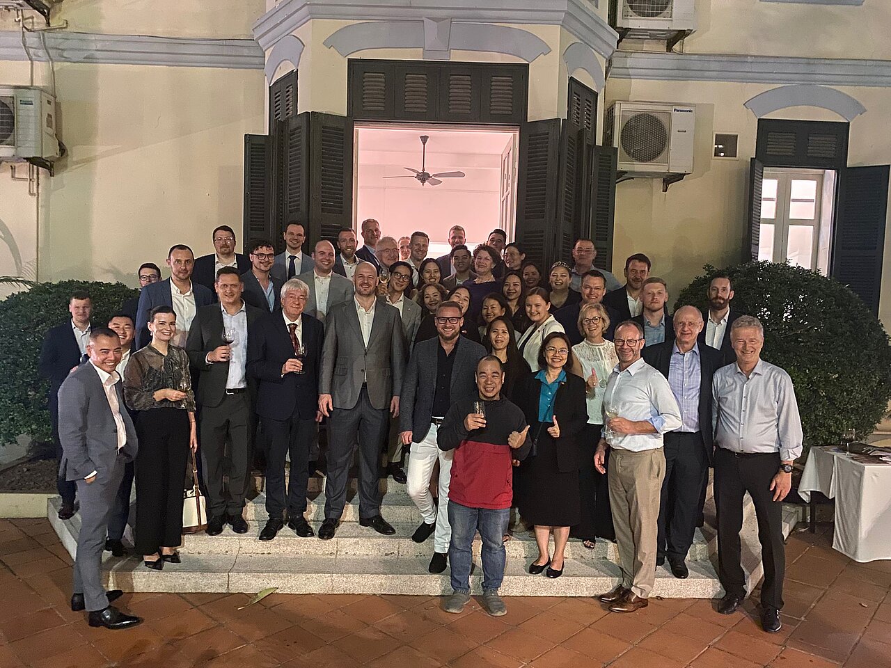 gruppenfoto-wirtschaftsdelegation-vietnam.jpg Gruppenfoto einer internationalen Wirtschaftsdelegation bei einer Abendveranstaltung in Vietnam vor einem historischen Gebäude.