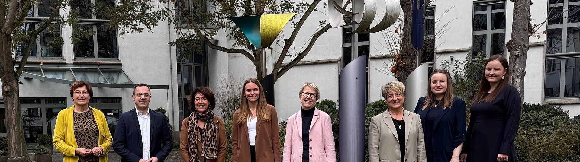 Eine Gruppe von neun Personen steht im Freien vor einem Gebäude und posiert für ein Foto. Hinter ihnen stehen mehrere Bäume sowie eine moderne Metallskulptur. Die Personen stehen nebeneinander auf einer Rasenfläche und lächeln in die Kamera.