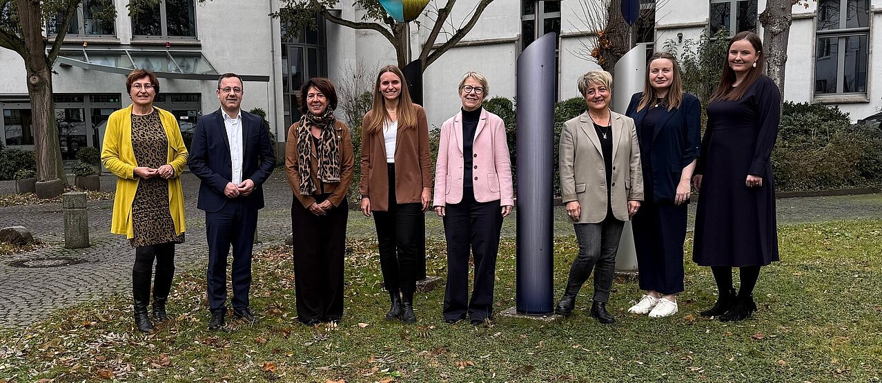 Eine Gruppe von neun Personen steht im Freien vor einem Gebäude und posiert für ein Foto. Hinter ihnen stehen mehrere Bäume sowie eine moderne Metallskulptur. Die Personen stehen nebeneinander auf einer Rasenfläche und lächeln in die Kamera.